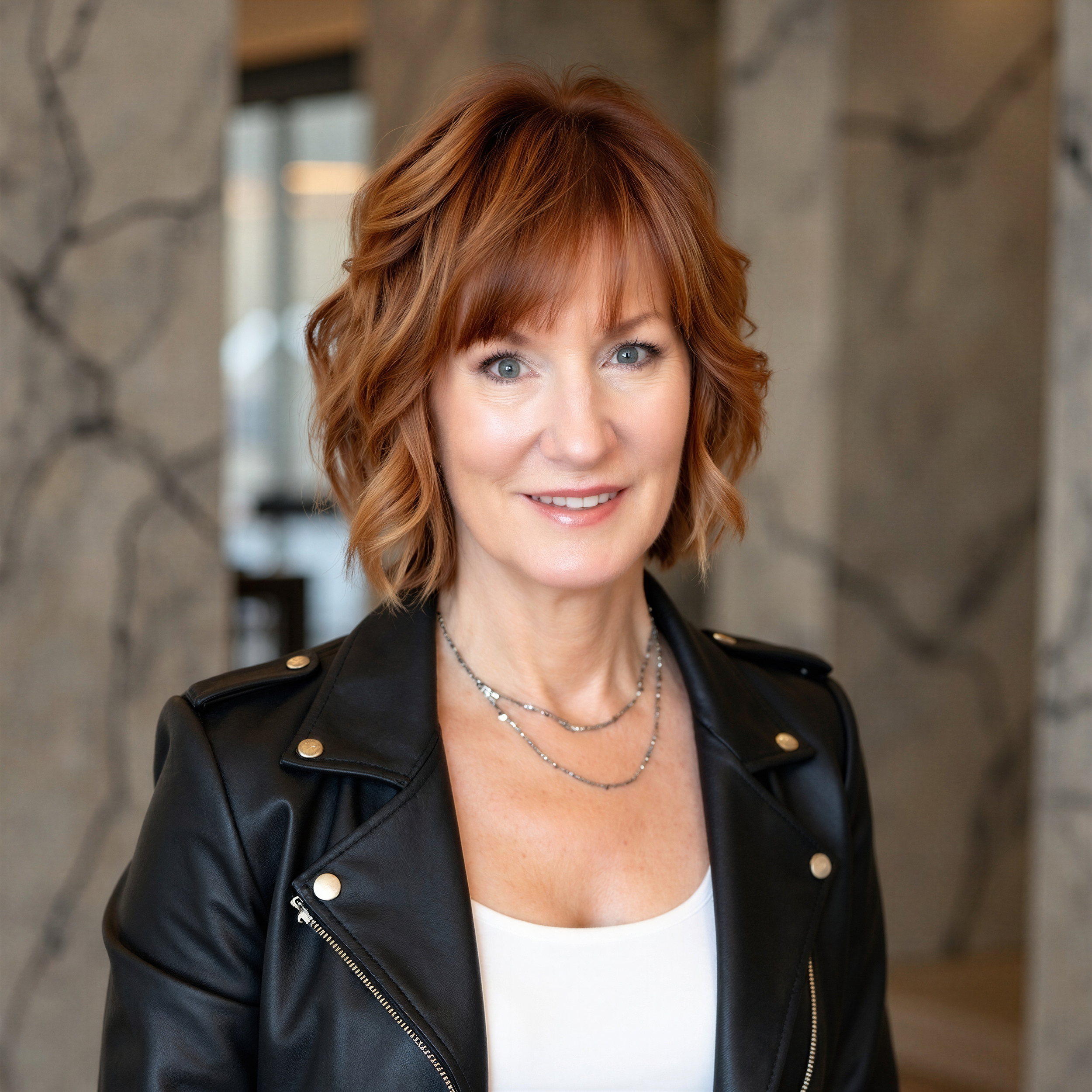 A woman with red hair styled in loose waves, wearing a black leather jacket with gold buttons over a white top, and layered necklaces. She is smiling and standing in front of a stone wall.