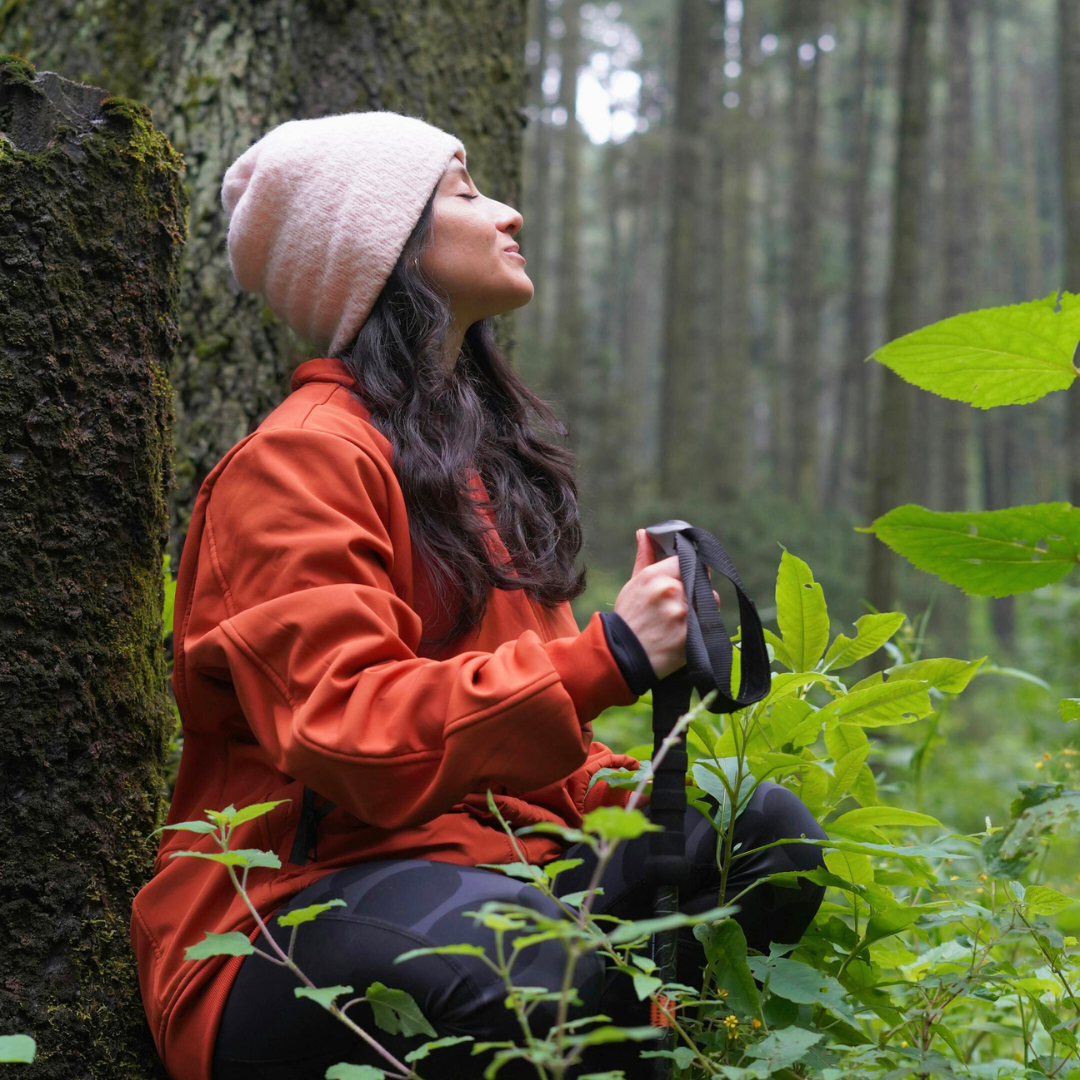 A woman in an orange jacket and pink beanie sits against a tree in a forest, holding a camera and looking peaceful with her eyes closed.