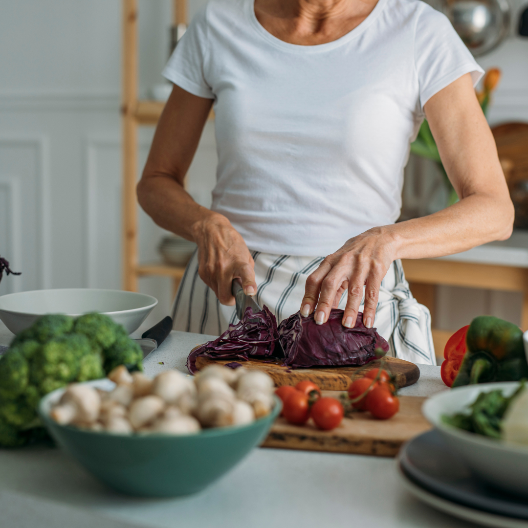 A person chopping red cabbage in a kitchen surrounded by fresh vegetables like broccoli, mushrooms, tomatoes, and peppers.