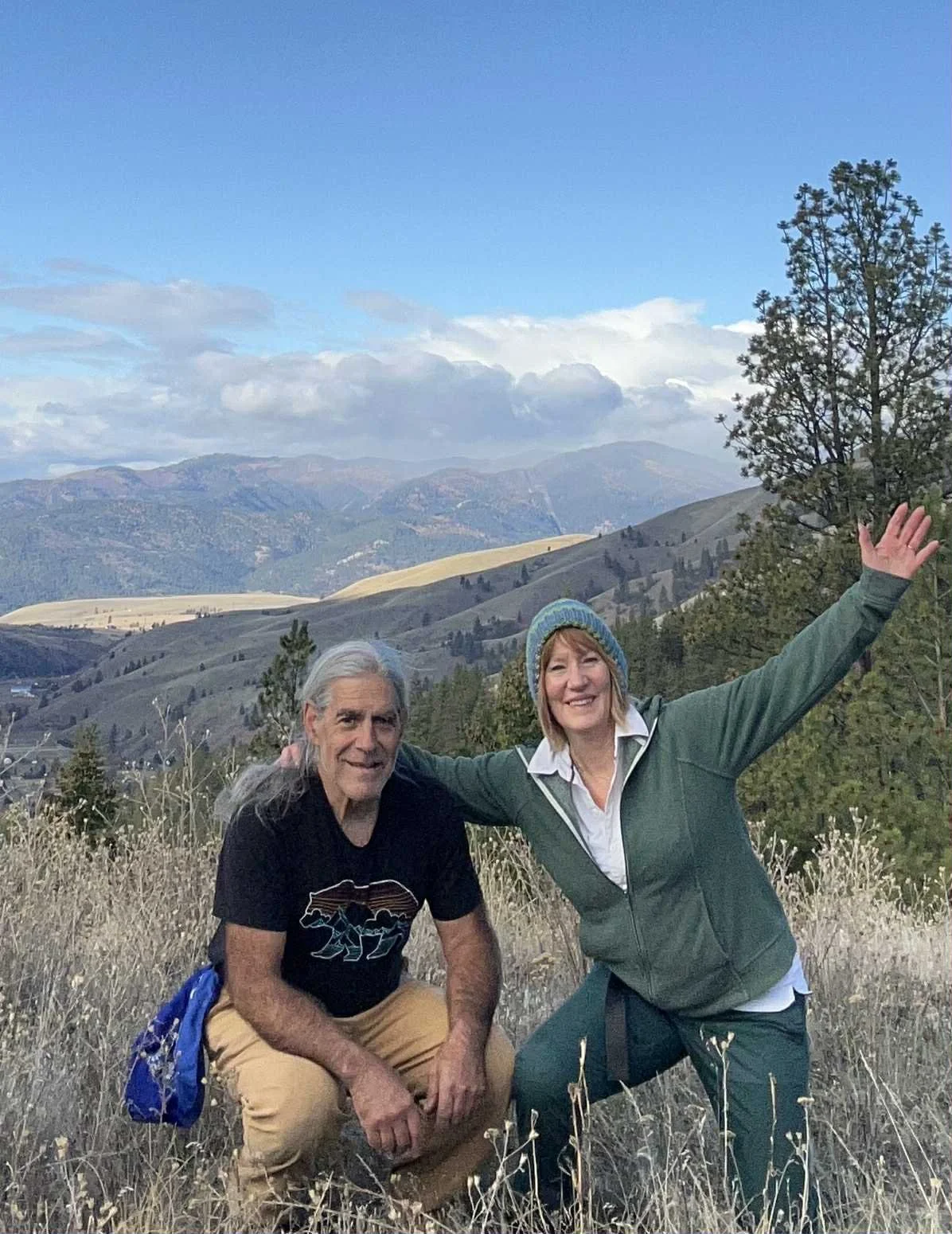 A man and woman hiking outdoors in a mountainous area with trees, dry grass, and a cloudy sky.