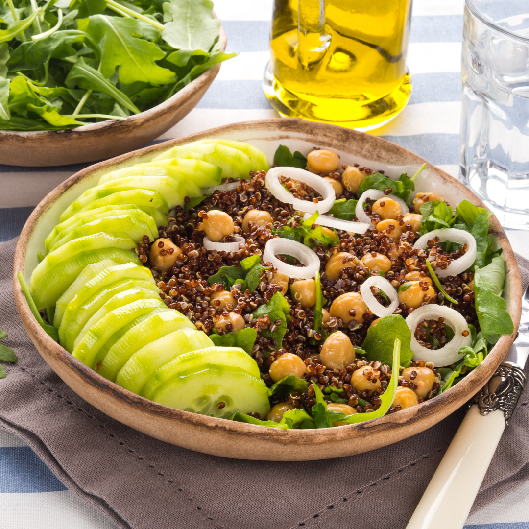 Fresh salad with black quinoa, chickpeas, cucumber slices, onion rings, and greens in a wooden bowl, with olive oil, water, and a side of greens on a table.