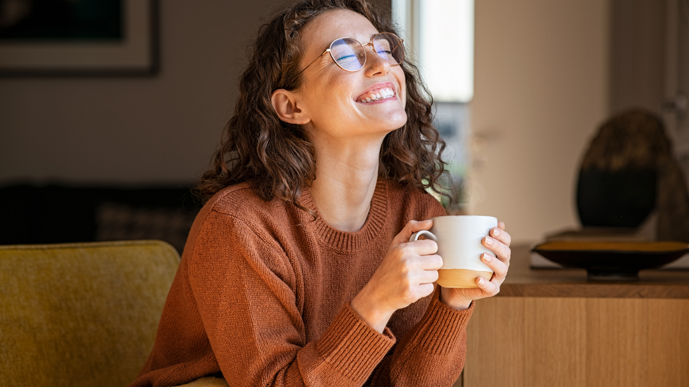 A woman with curly brown hair and glasses smiling with her eyes closed, holding a tan and white mug, sitting in a cozy room.