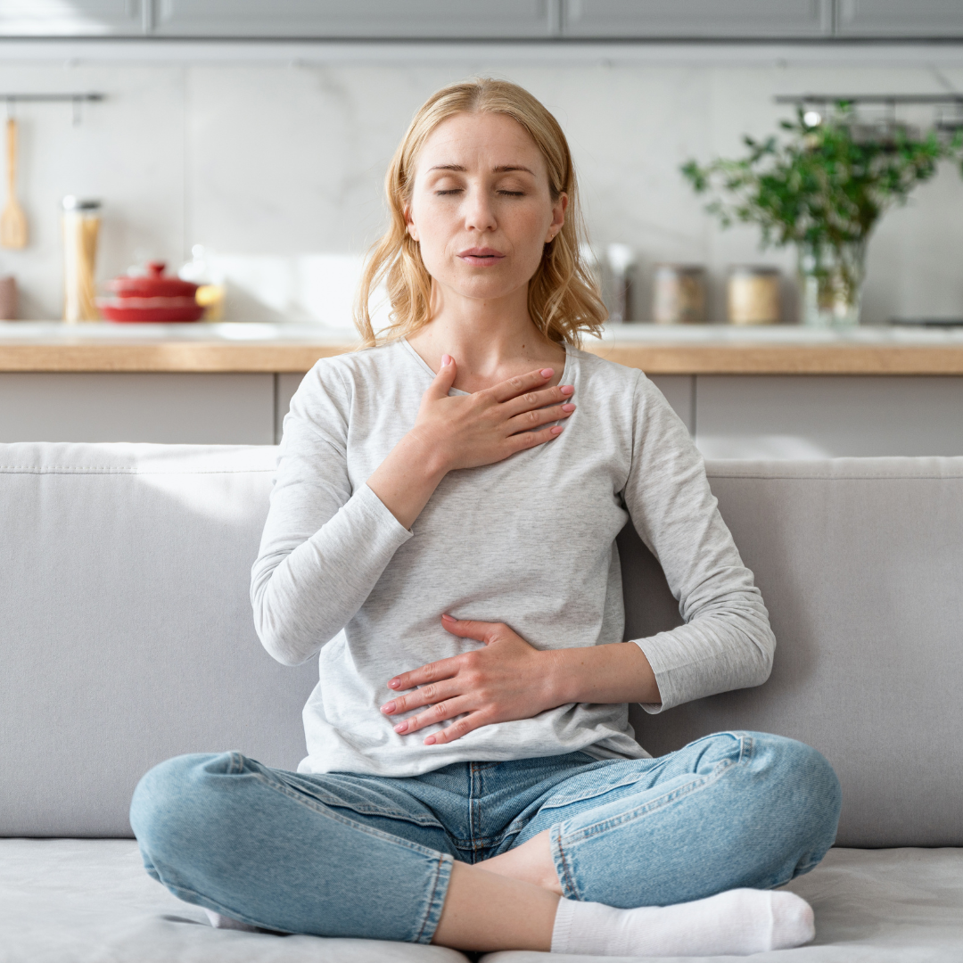 A woman sitting cross-legged on a couch with one hand on her chest and the other on her stomach, appearing to be experiencing discomfort or nausea in a modern kitchen setting.