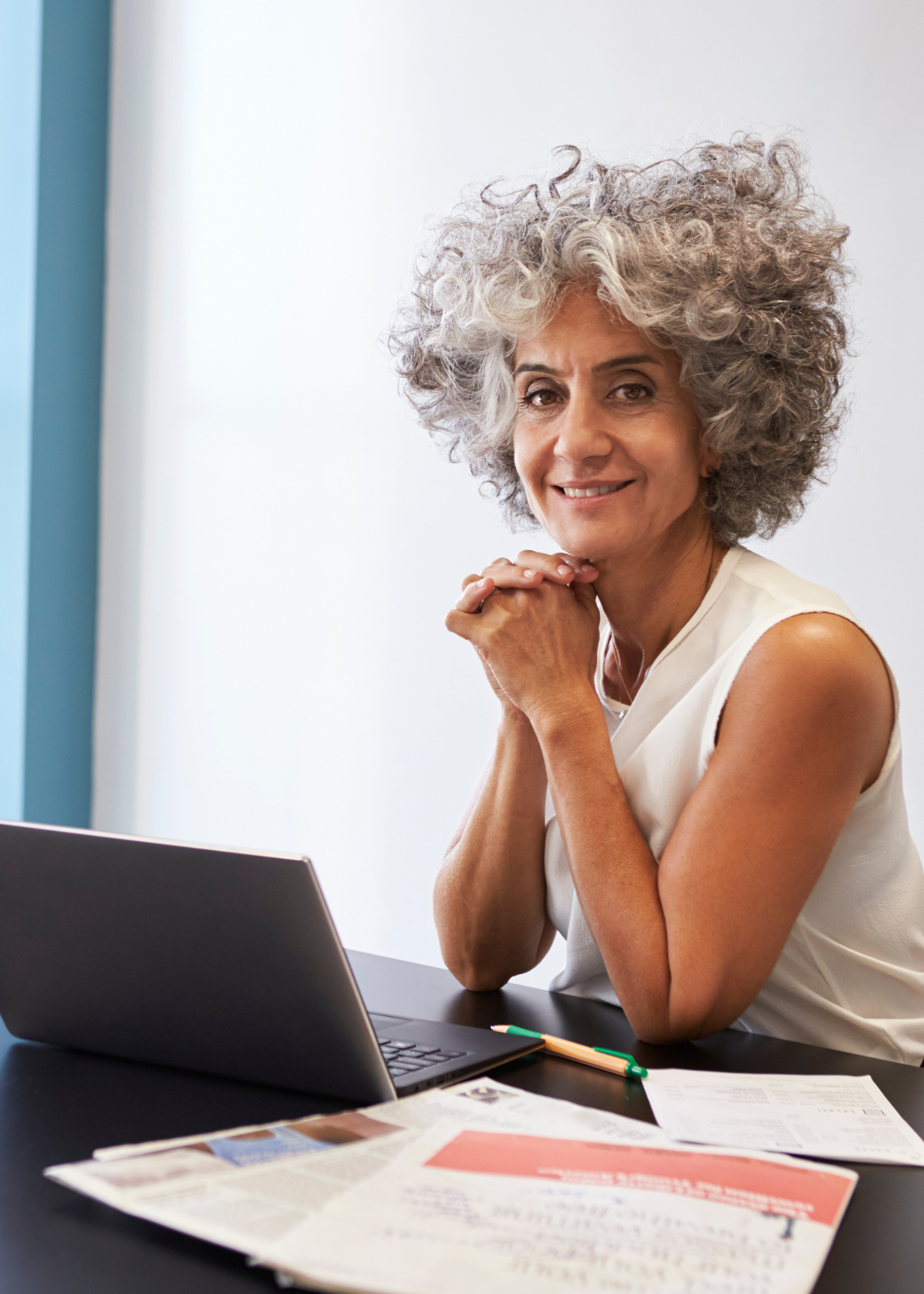 A smiling elderly woman with curly gray hair sitting at a desk with a laptop, papers, and a pen in front of her, looking at the camera.