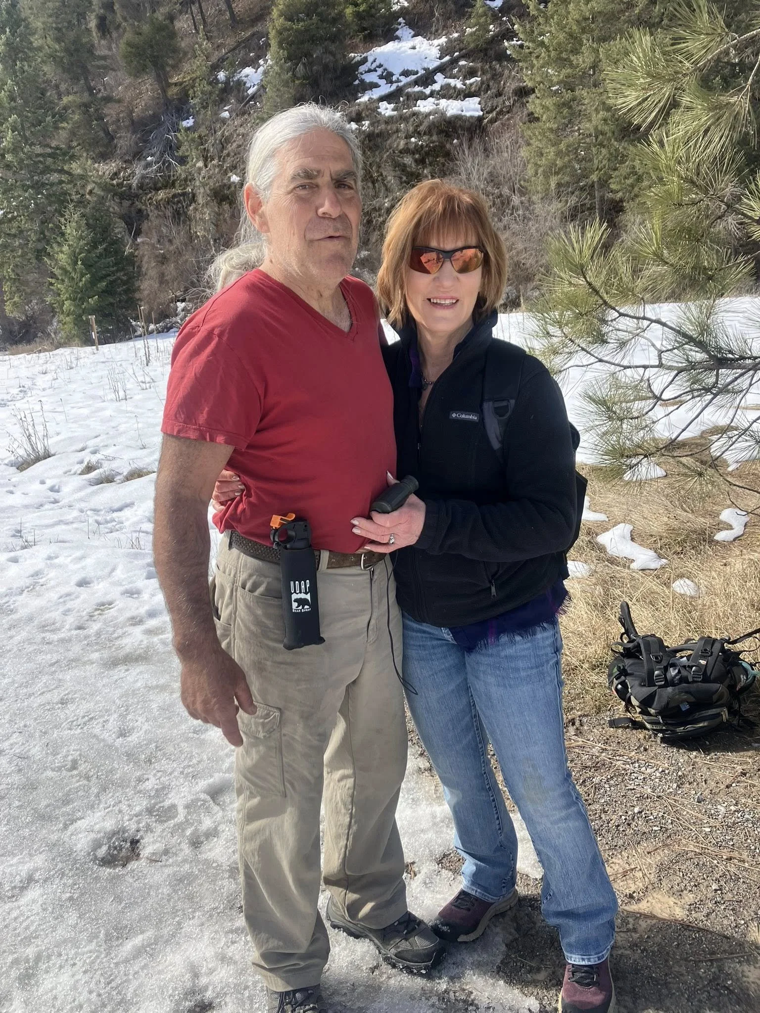 A man in a red t-shirt and beige pants standing next to a woman in a black jacket and blue jeans on a snowy mountain trail with trees around.