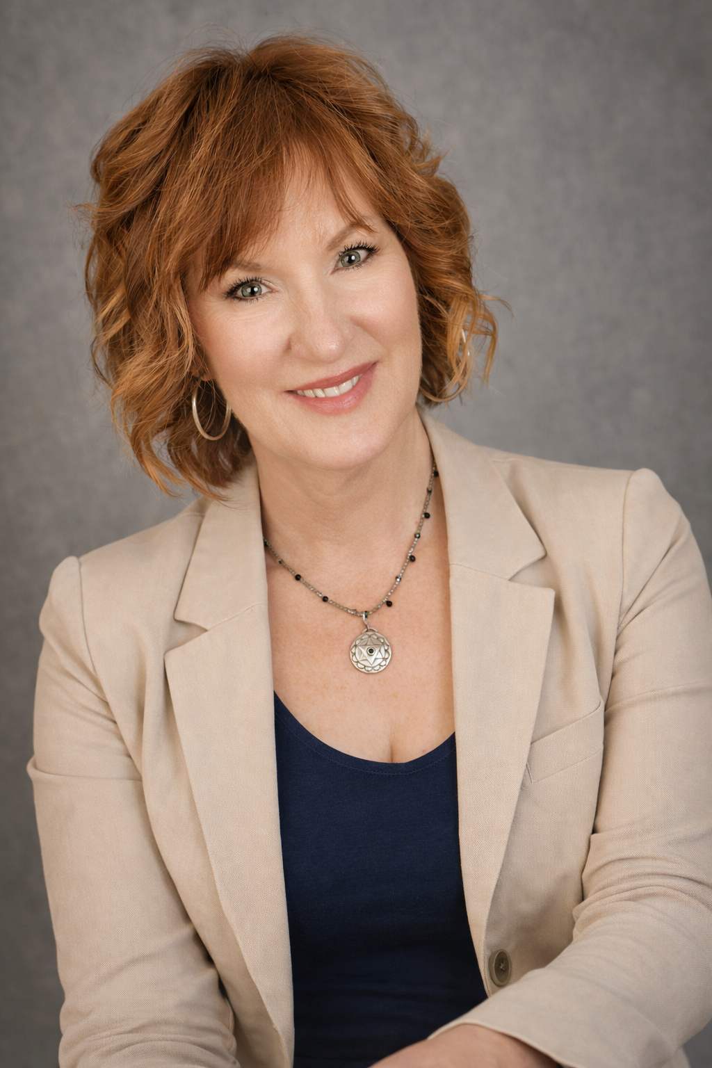 A woman with curly red hair wearing a beige blazer, dark blue top, hoop earrings, and a round pendant necklace, smiling at the camera against a gray background.