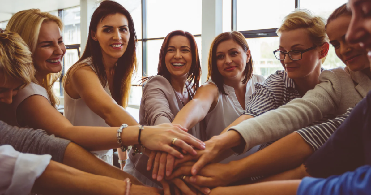 Group of women in casual clothing putting their hands together in a show of unity and teamwork in a brightly lit room with large windows.