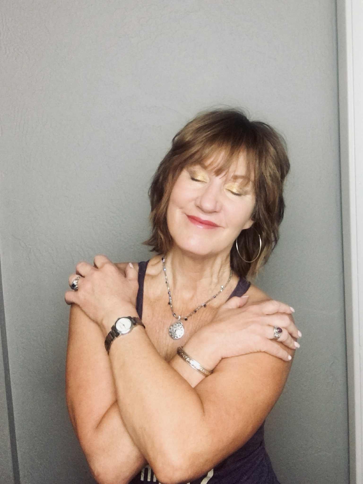 A woman with short brown hair and closed eyes, smiling with her arms crossed over her shoulders, standing against a plain gray wall.