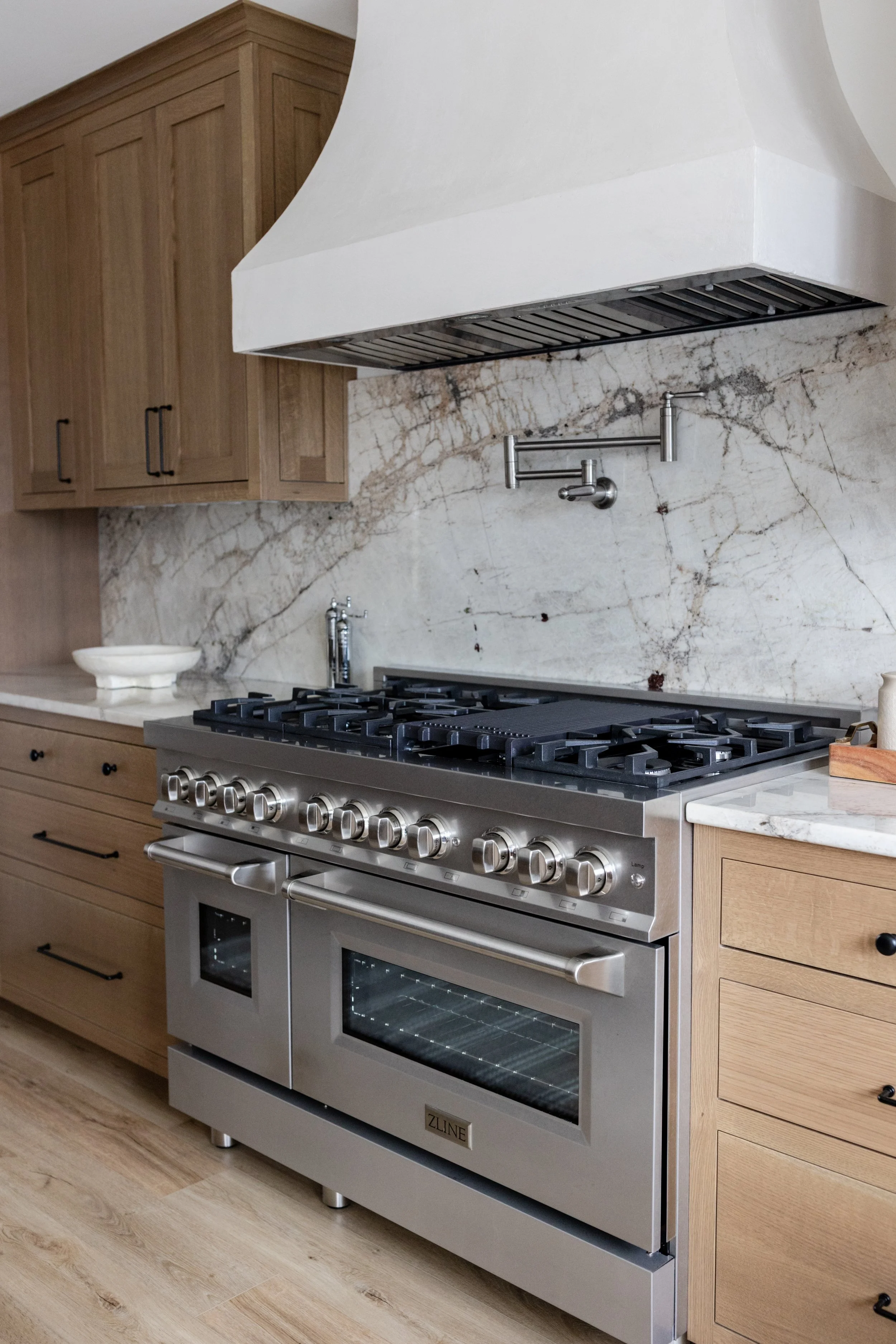 A modern kitchen featuring a stainless steel ZLINE range oven with a gas cooktop, wooden cabinetry, a marble backsplash, and a white range hood.