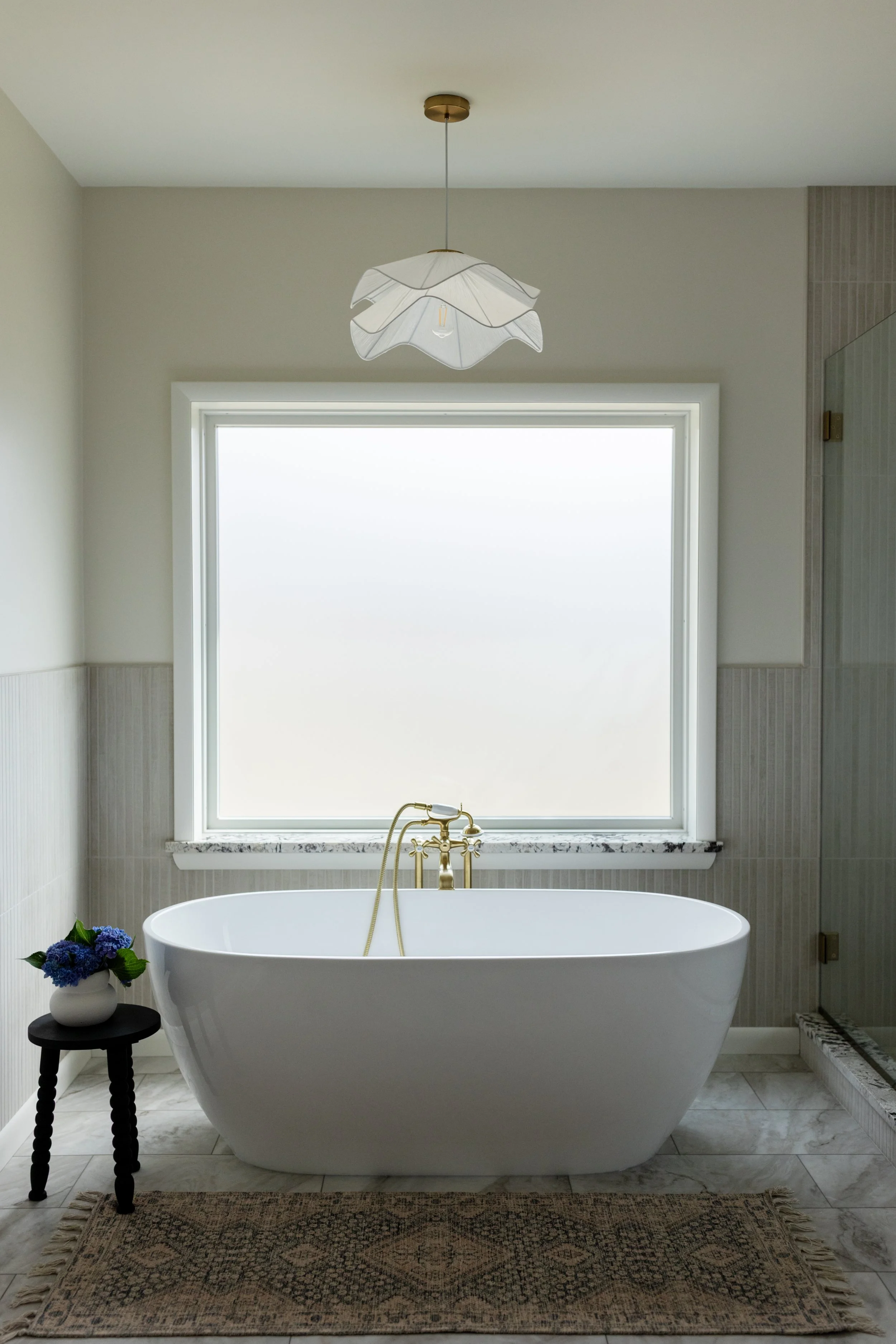 Elegant bathroom with a white freestanding bathtub, a black side table with a vase of purple flowers, ornate rug, large frosted window, brass faucet, and modern light fixture.