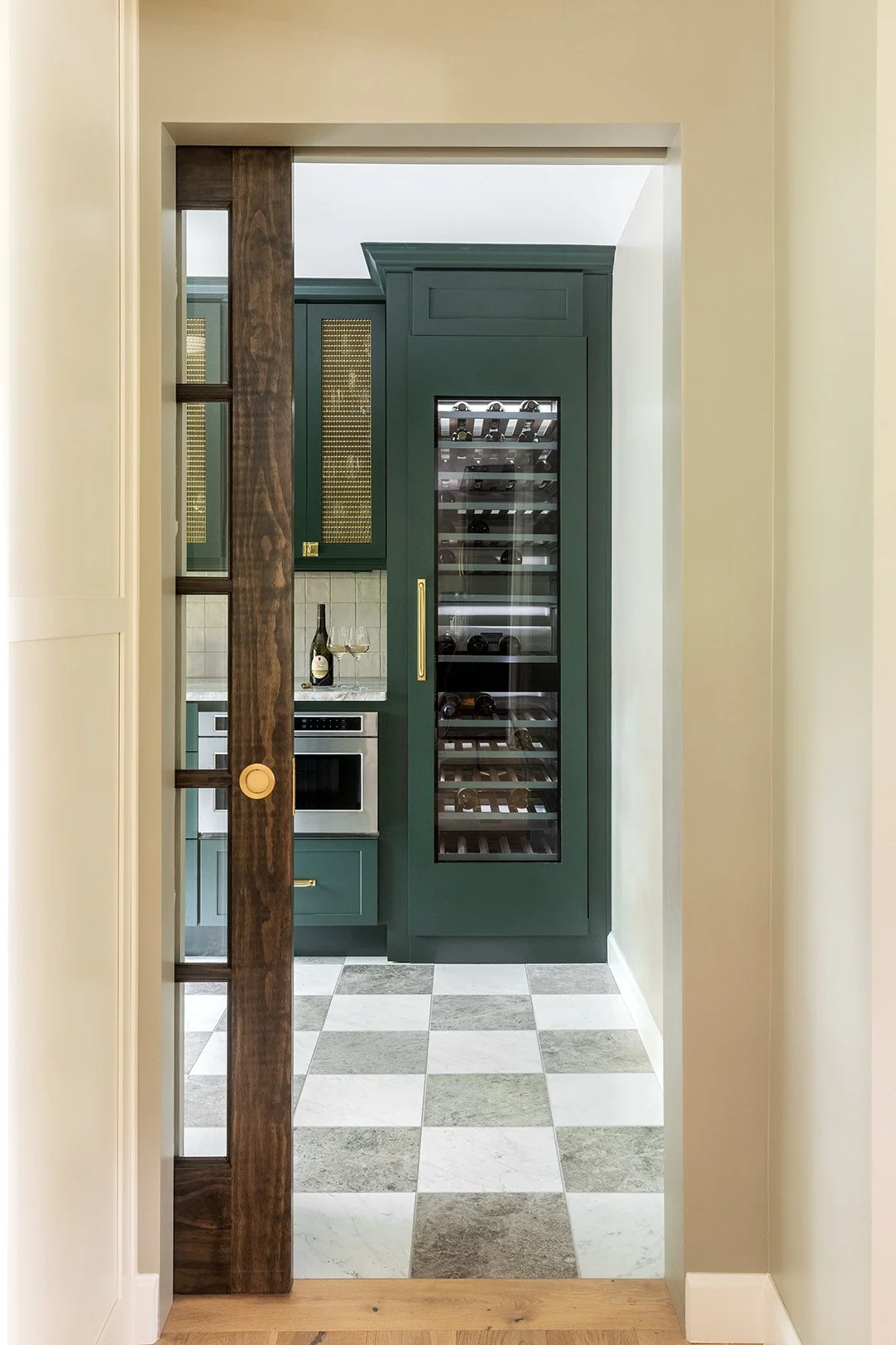 A view through a doorway into a kitchen with green cabinets, a wine refrigerator, a wine bottle, and two wine glasses on the counter. The floor has a checkered pattern of white and gray tiles.