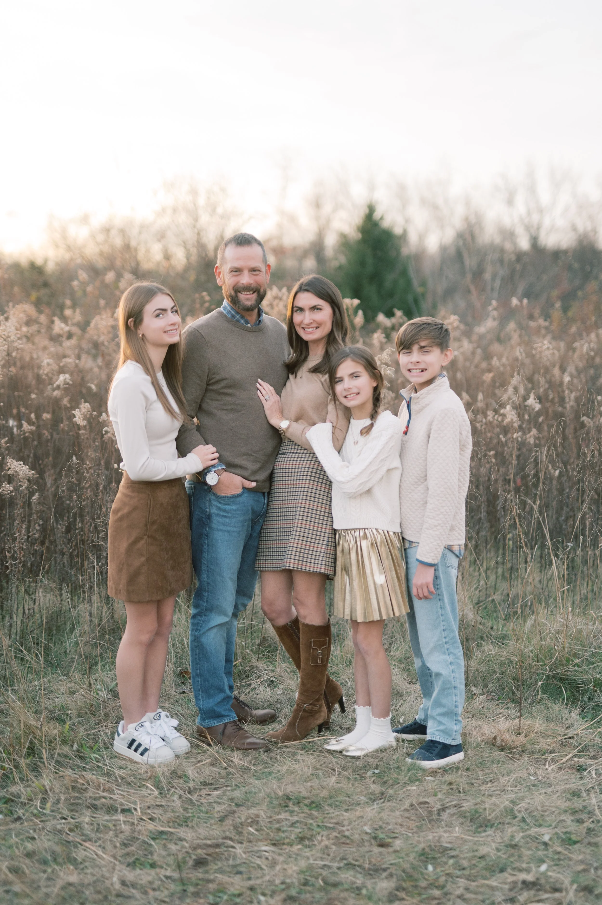 A family of five standing outdoors in a field with tall grass and trees in the background during golden hour. They are smiling and dressed in casual fall clothing.
