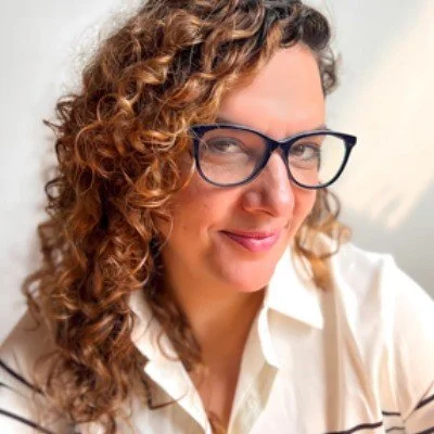 A woman with curly hair wearing glasses and a white shirt, smiling at the camera.