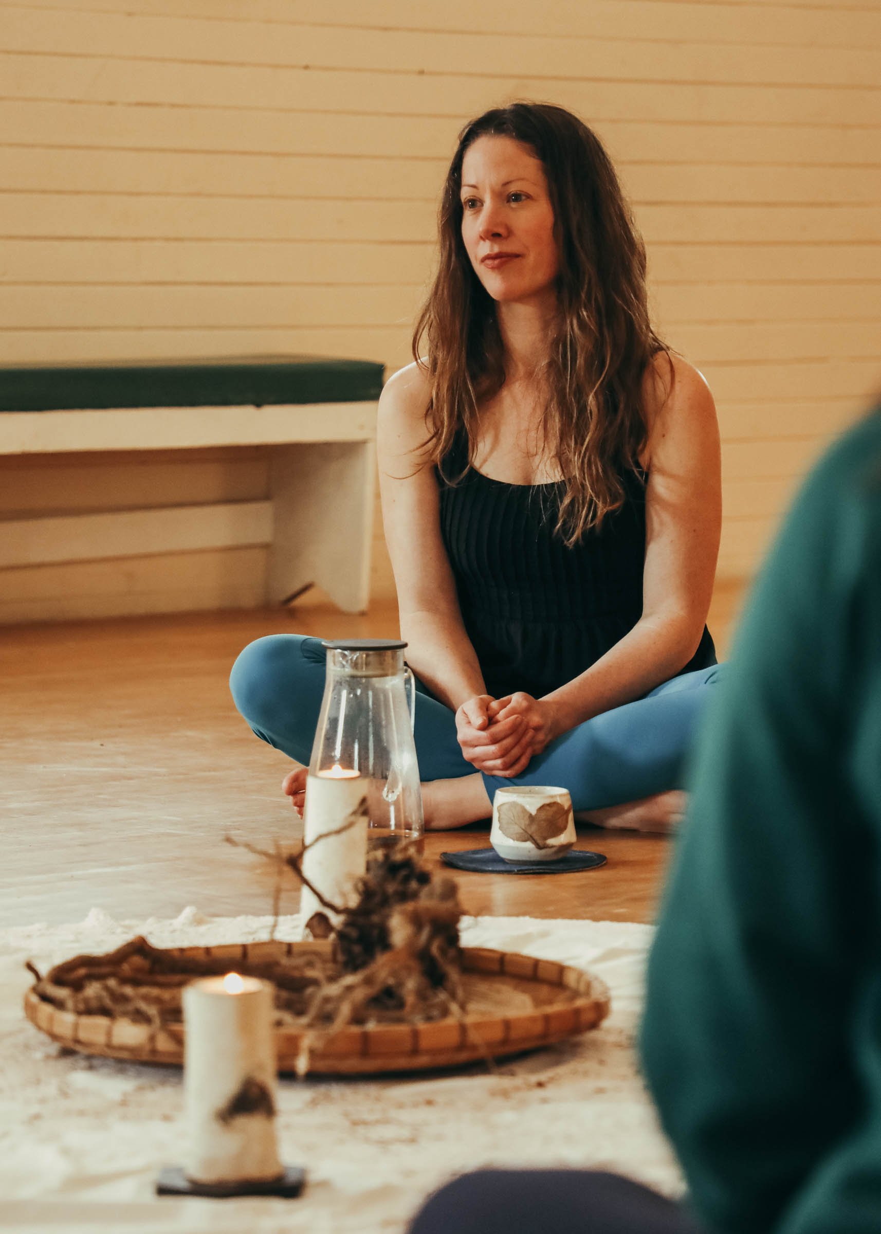 A woman with long brown hair sitting cross-legged on the floor, wearing a black sleeveless top and blue pants, with candles and decorative items on a low wooden table in front of her, and a light wooden wall in the background.