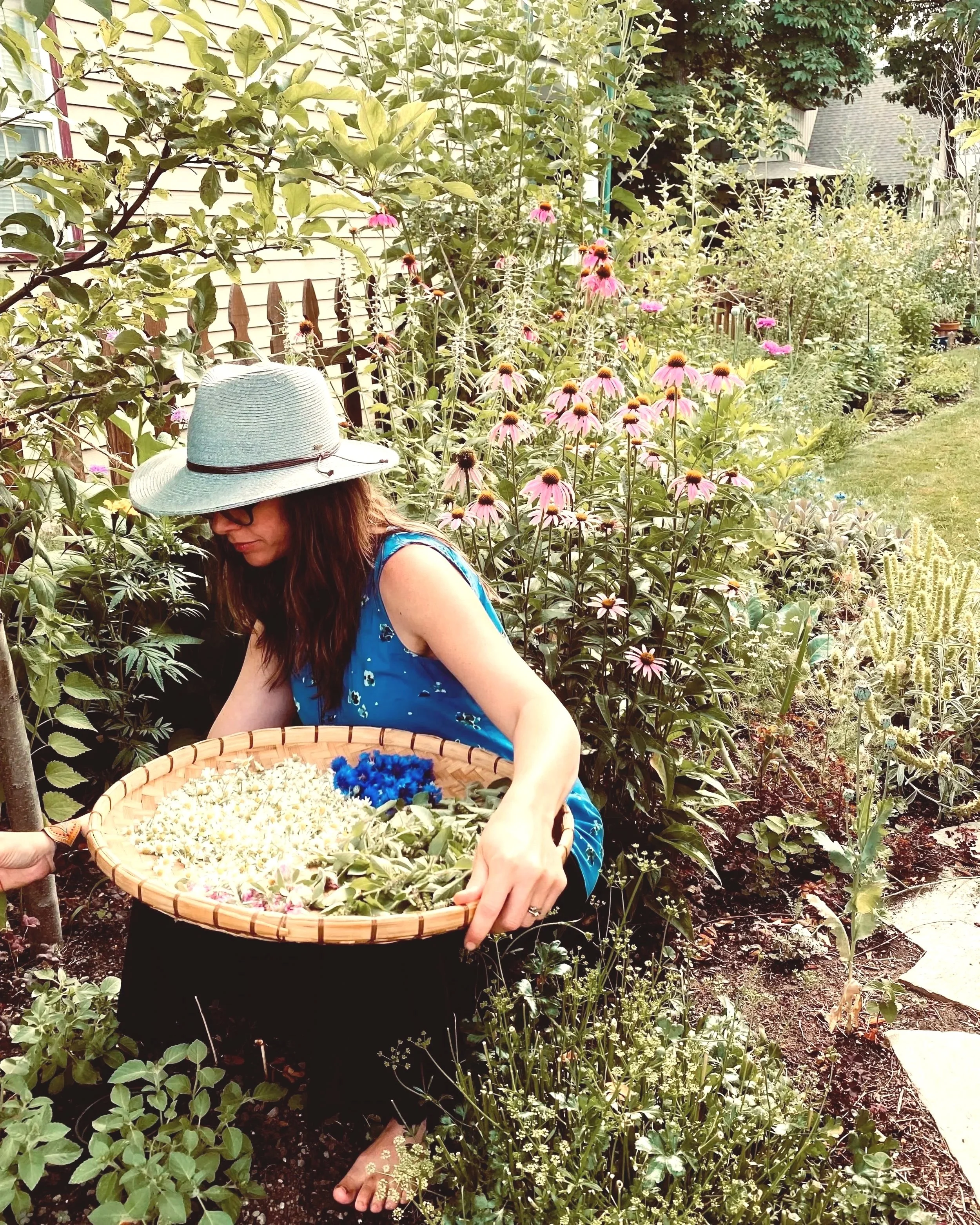 A woman wearing a light blue dress, black glasses, and a wide-brimmed hat is in a garden, holding a large basket full of white and blue flowers, surrounded by various flowering plants and greenery.