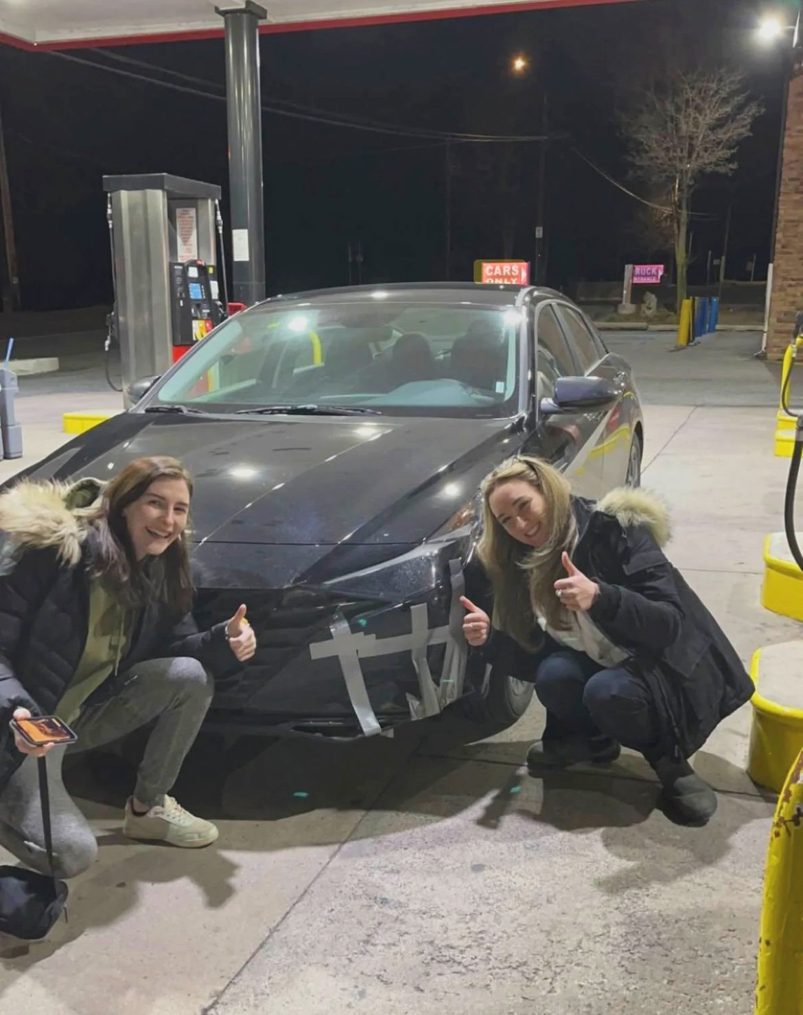 Two girls giving a thumbs up beside a car with duct tape on the bumper