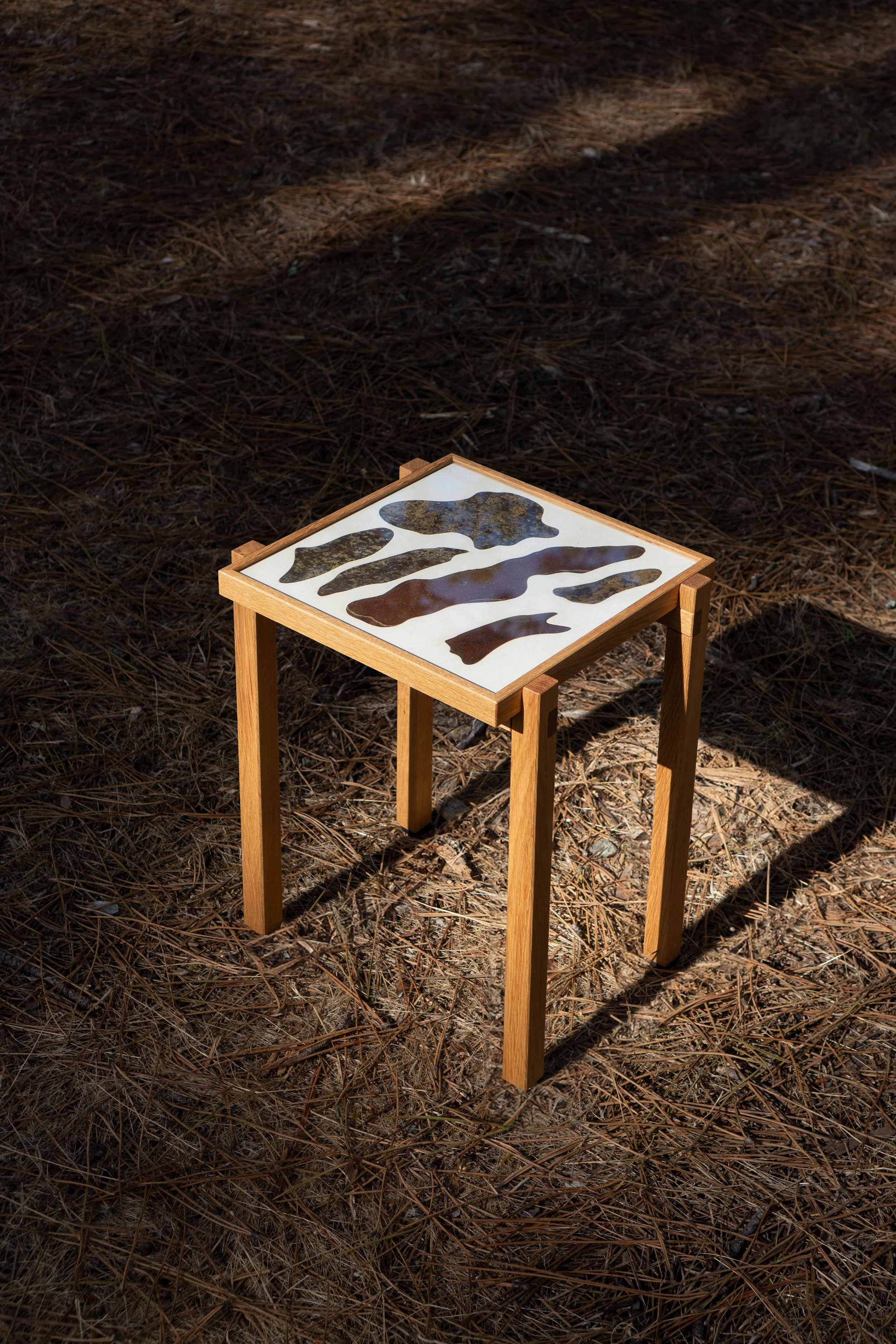 A small wooden table outdoors on a ground covered with pine needles. The table top displays an abstract pattern with irregular, wavy shapes in various dark colors.