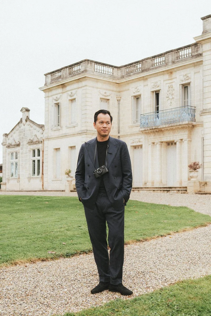Homme en costume noir avec un appareil photo autour du cou, debout devant un bâtiment en pierre blanche avec un petit jardin.