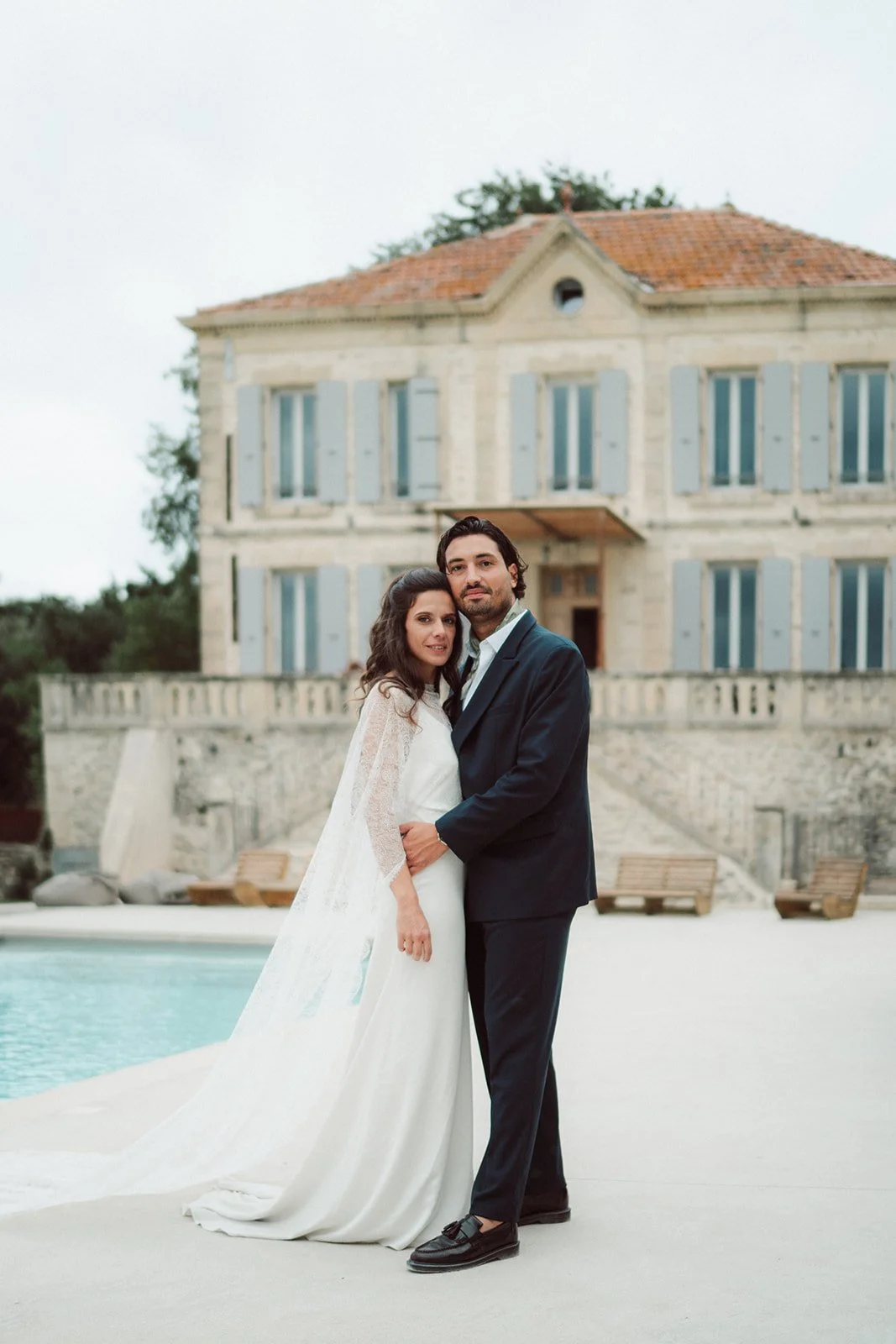 Un couple en tenue de mariage debout près d'une piscine devant une grande maison ancienne avec volets bleus.