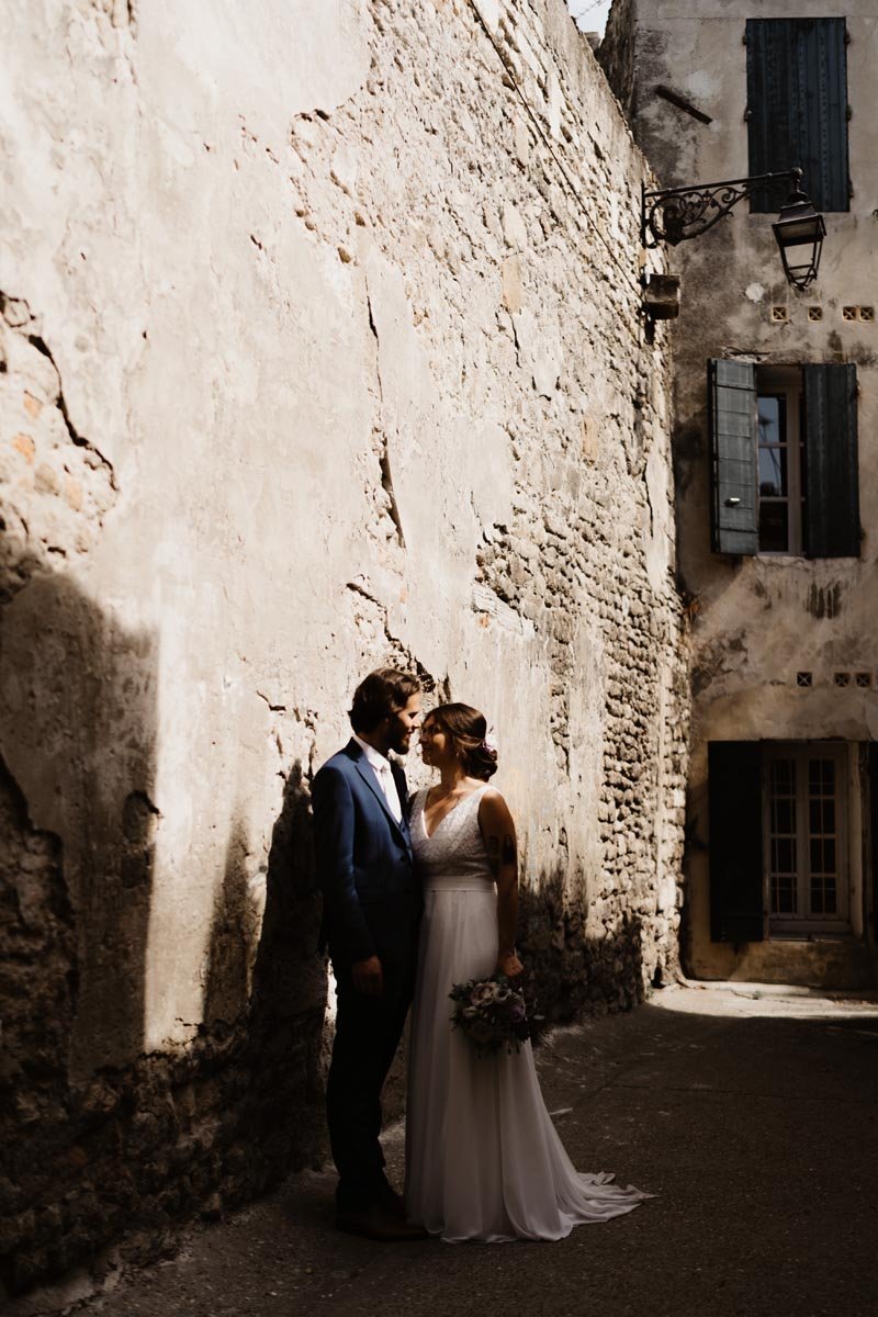 Un couple en tenue de mariage se tient proche dans une ruelle étroite avec des murs en pierre, éclairée par un lampadaire.