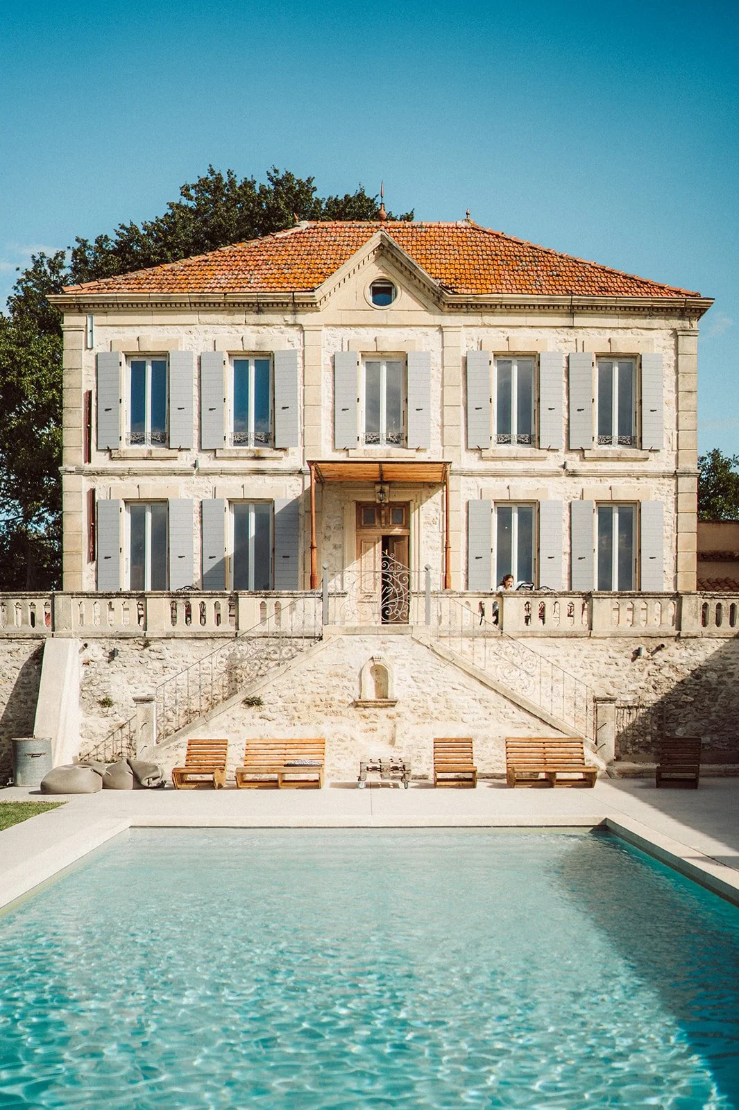 Maison ancienne à trois étages avec volets bleus, face à une piscine, sous un ciel bleu.