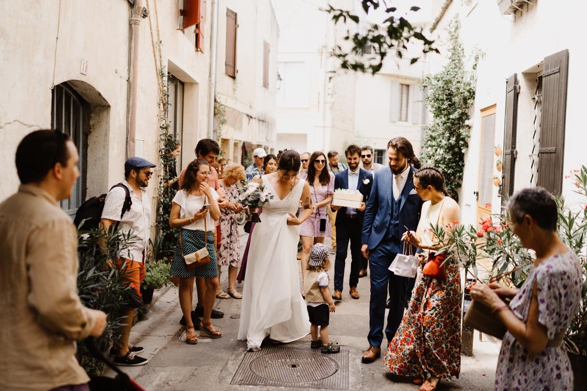 Un groupe de personnes dans une rue, avec une mariée en robe blanche et un marié en costume bleu entourés d'invités. Un enfant se tient devant eux.