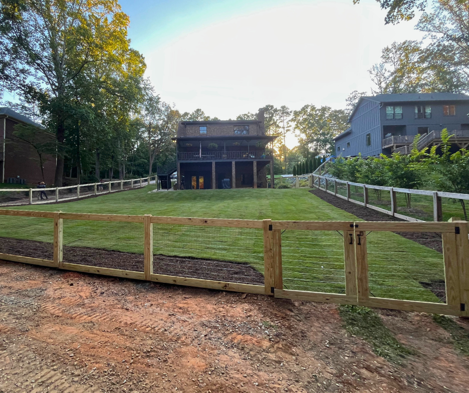 Large backyard with freshly installed sod enclosed by a new wooden post-and-rail fence, surrounding a two-story home at the top of the hill with mature trees on the left and a neighbouring home on the right