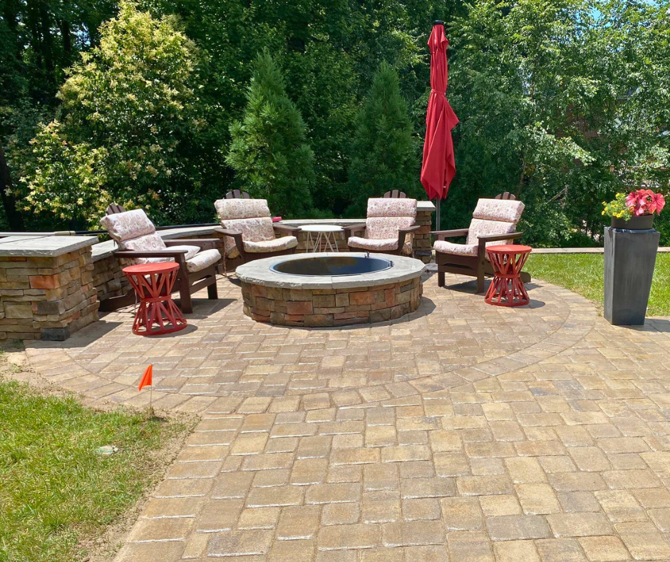 Outdoor living space with a circular stacked-stone fire pit centered on a curved paver patio, surrounded by cushioned chairs, decorative red tables, and a tall red umbrella against a background of lush trees