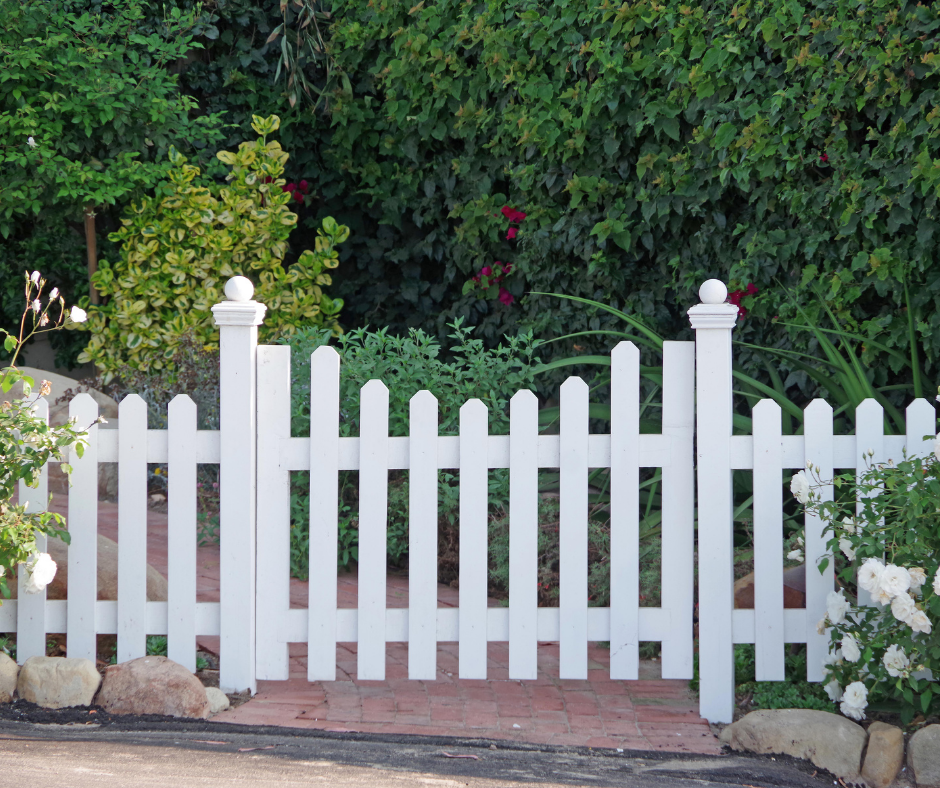 White picket fence with decorative posts surrounding a garden walkway lined with shrubs and flowering plants.