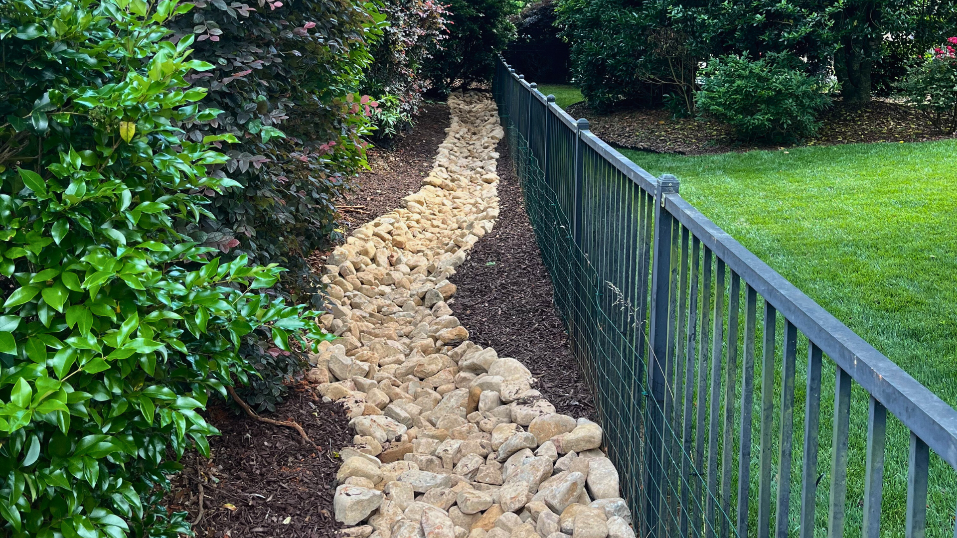 Stone-lined drainage channel running between dense shrubs and a black metal fence in a landscaped backyard, surrounded by fresh brown mulch.