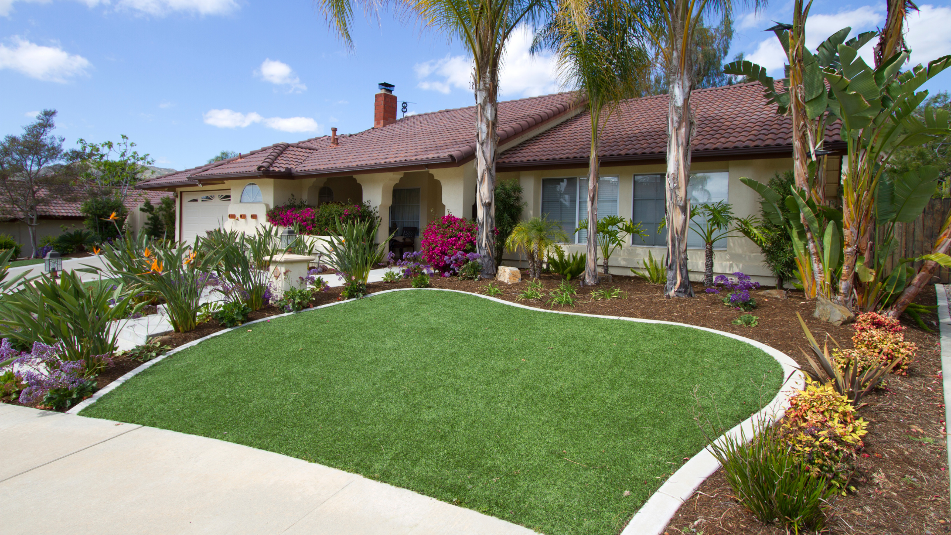 Front yard with artificial turf, palm trees, flowering shrubs, bird of paradise plants, and mulch beds in front of a single-story off white home with a tile roof