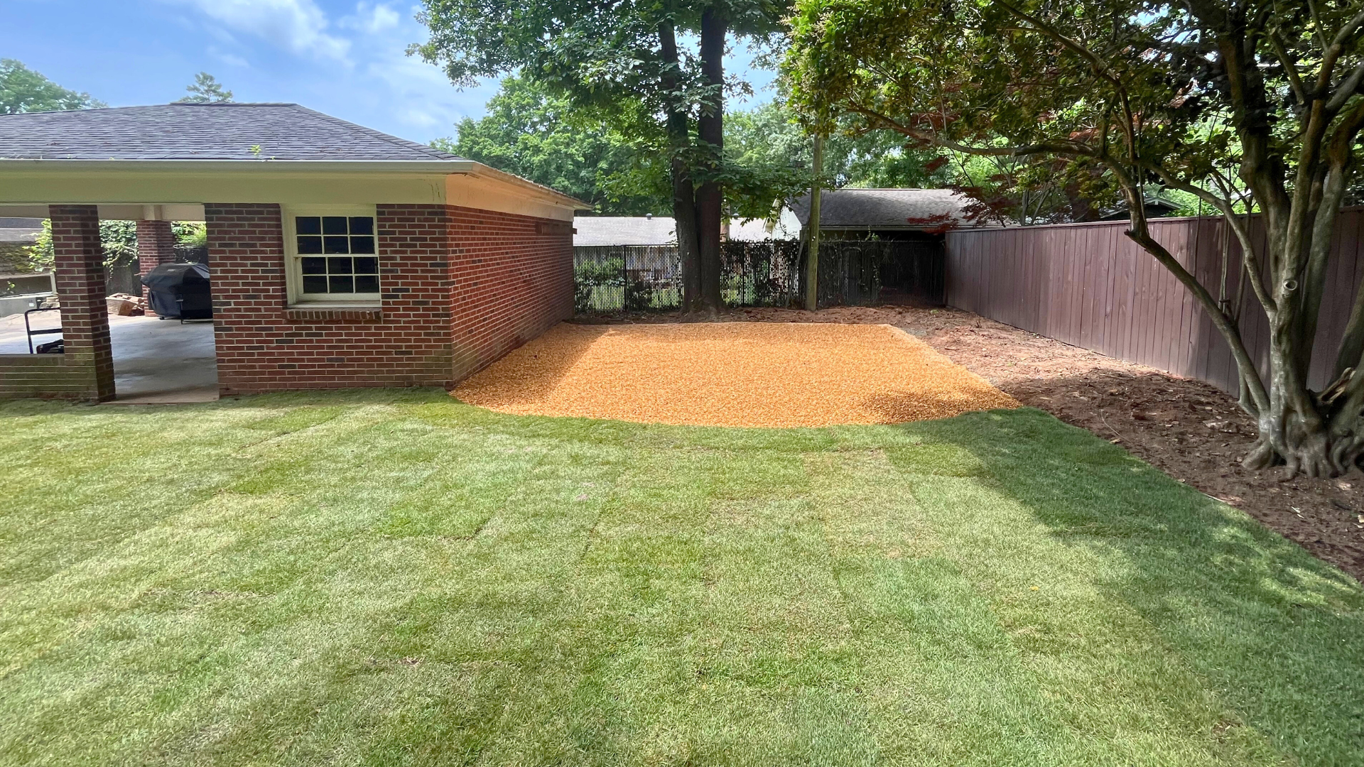Landscape renovation featuring freshly laid sod and a graded gravel area next to a brick outbuilding, with trees and a wooden privacy fence along the property line.