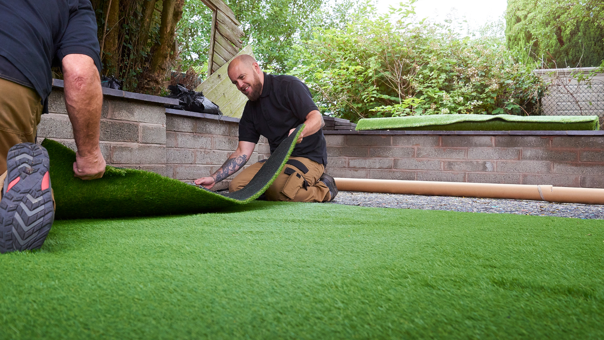 Two workers installing artificial turf in a landscaped yard, unrolling and trimming synthetic grass on a leveled surface beside stone retaining walls and greenery.