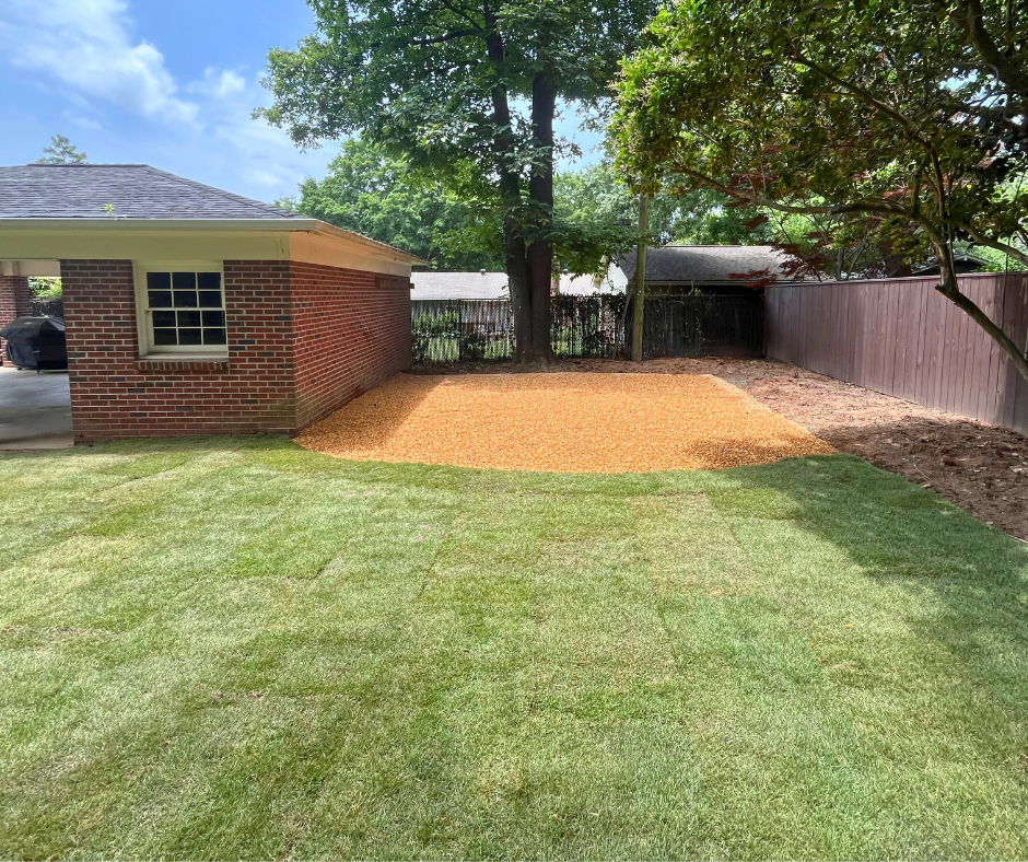 Landscape renovation featuring freshly laid sod and a graded gravel area next to a brick outbuilding, with trees and a wooden privacy fence along the property line