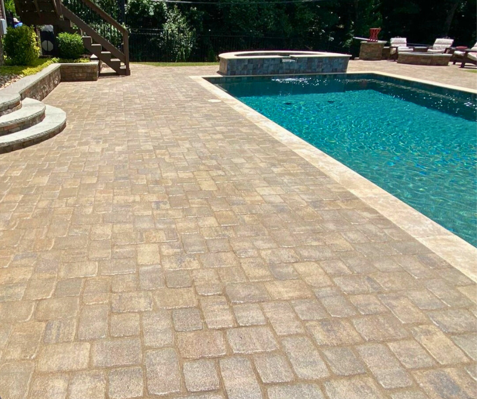Backyard pool area featuring a spacious stone paver patio, raised seating wall, and curved entry steps beside a blue inground pool