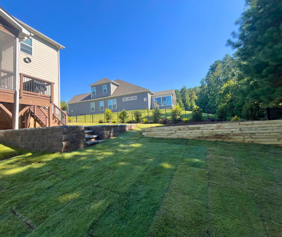 Residential backyard with newly installed sod, tiered wooden retaining walls, and stone steps leading to an elevated landscaped garden bed area with small green shrubs and fresh brown mulch
