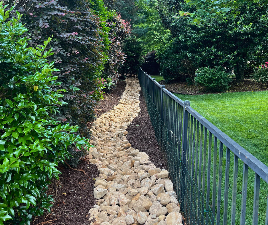 Stone-lined drainage channel running between dense shrubs and a black metal fence in a landscaped backyard, surrounded by fresh brown mulch