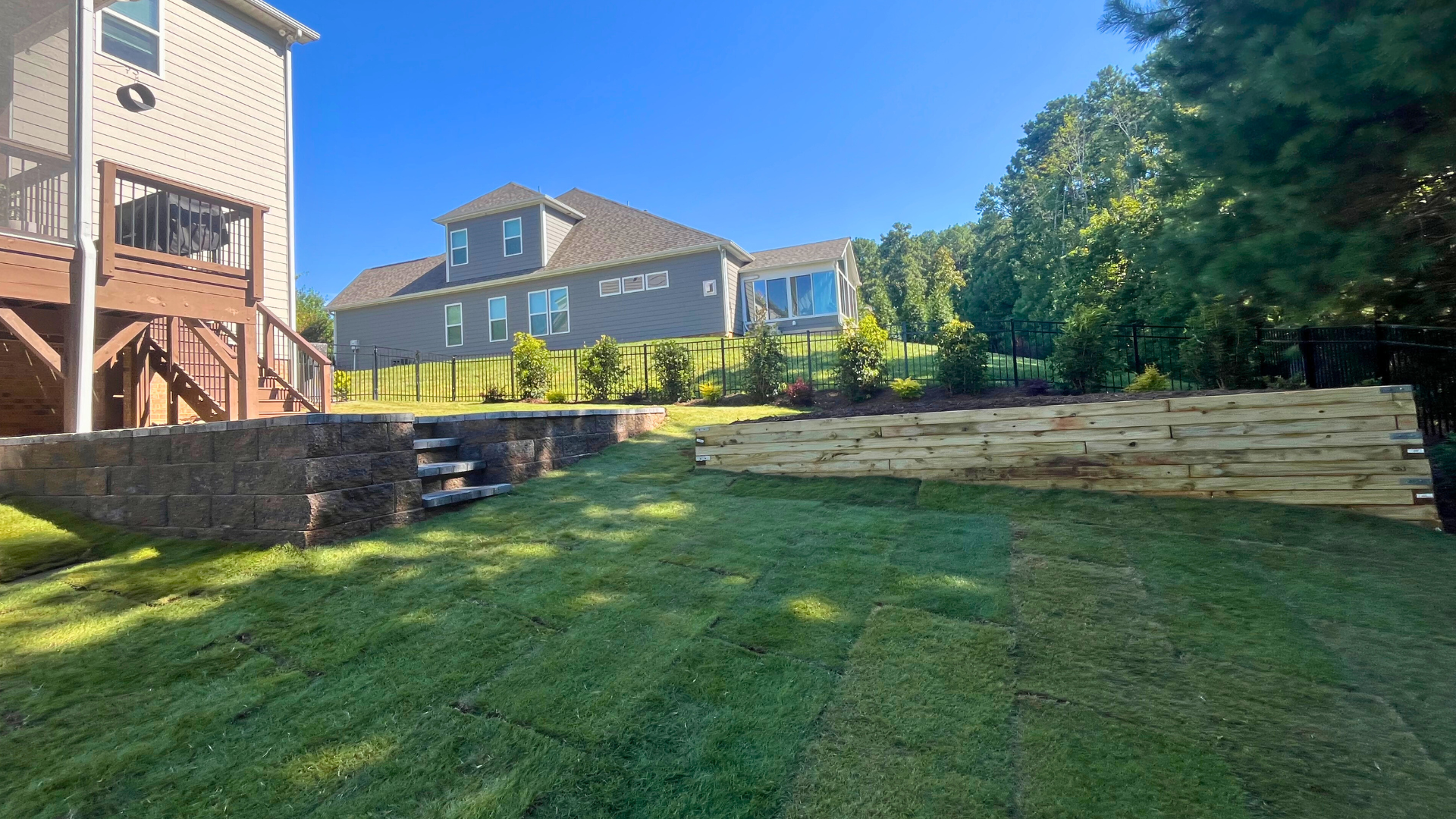 Residential backyard with newly installed sod, tiered wooden retaining walls, and stone steps leading to an elevated landscaped garden bed area with small green shrubs and fresh brown mulch.