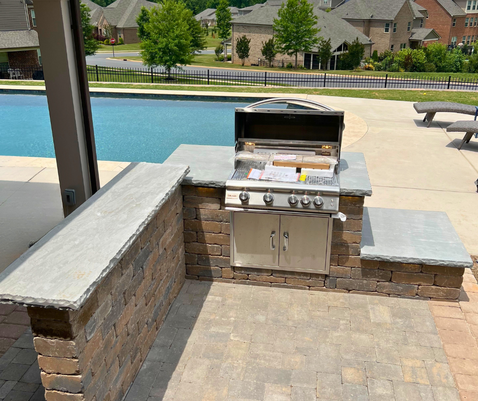 Poolside outdoor kitchen featuring a stainless steel built-in grill integrated into a stone island with concrete slab countertops, set on a paver patio in a suburban backyard