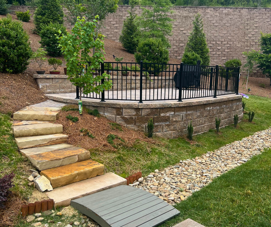 Tiered backyard with stone slab steps leading to a raised paver patio enclosed by a black metal railing, alongside a dry creek bed lined with river rock.