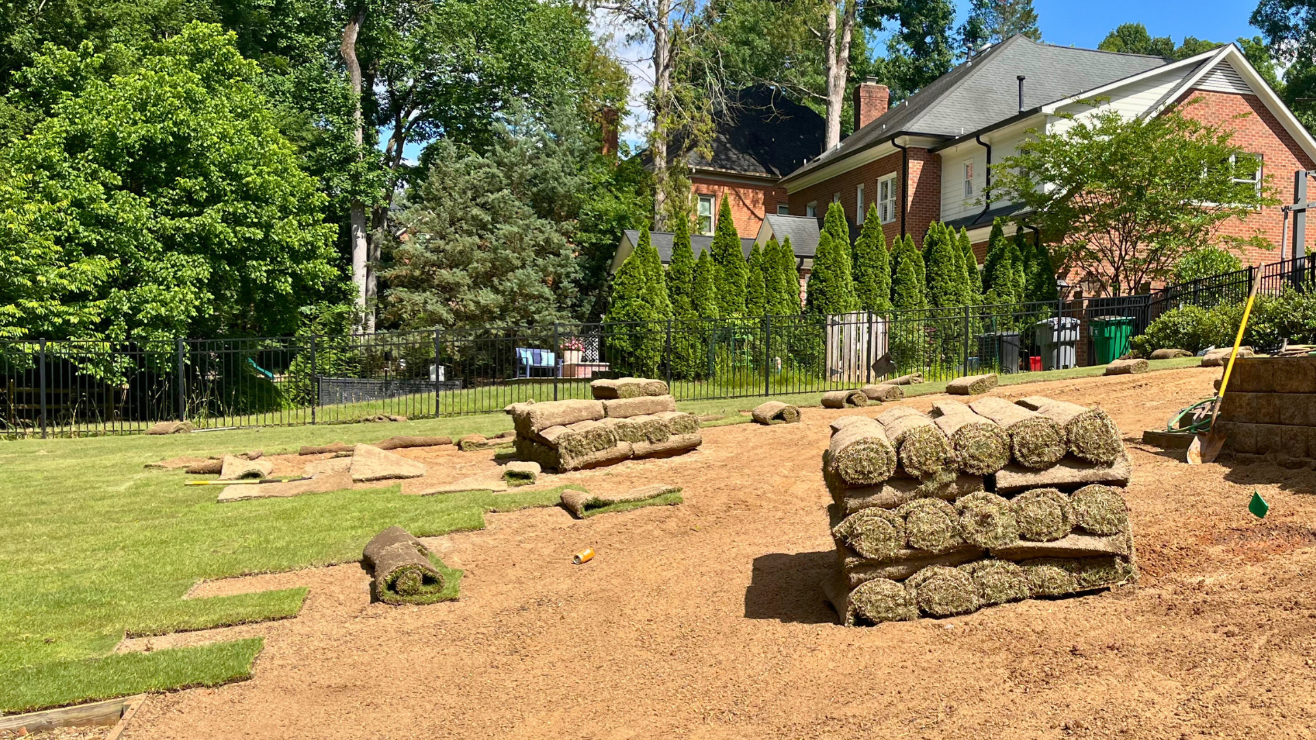 Sod installation in progress with piles of rolled sod ready for installation on freshly graded soil, with partially laid green sod, surrounded by mature trees, thin black fencing, and neighbouring homes
