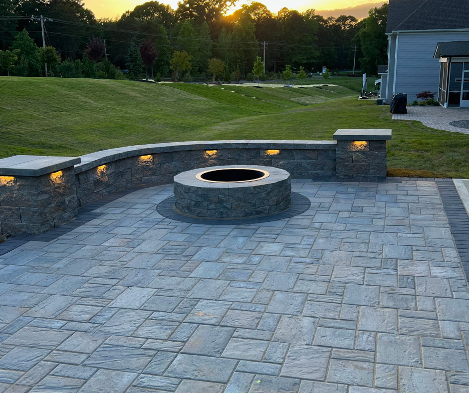 Stone backyard patio with a circular fire pit and low retaining wall featuring built-in landscape lighting at sunset.