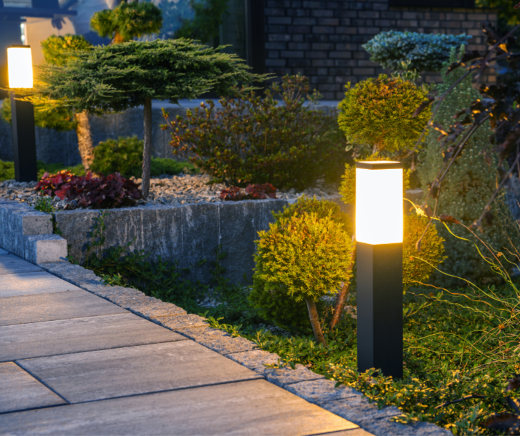 Sleek black metal bollard lights with illuminated frosted glass tops lining a stone pathway, providing soft, warm lighting over manicured shrubs and a decorative garden at dusk.