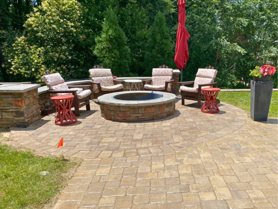 Outdoor living space with a circular stacked-stone fire pit centered on a curved paver patio, surrounded by cushioned chairs, decorative red tables, and a tall red umbrella against a background of lush trees.