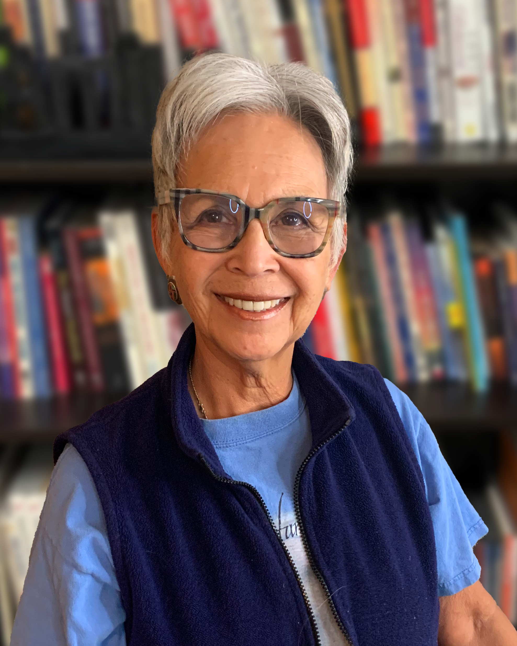 Smiling elderly woman with short gray hair, glasses, wearing a blue vest over a light blue shirt, standing in front of bookshelves filled with colorful books.