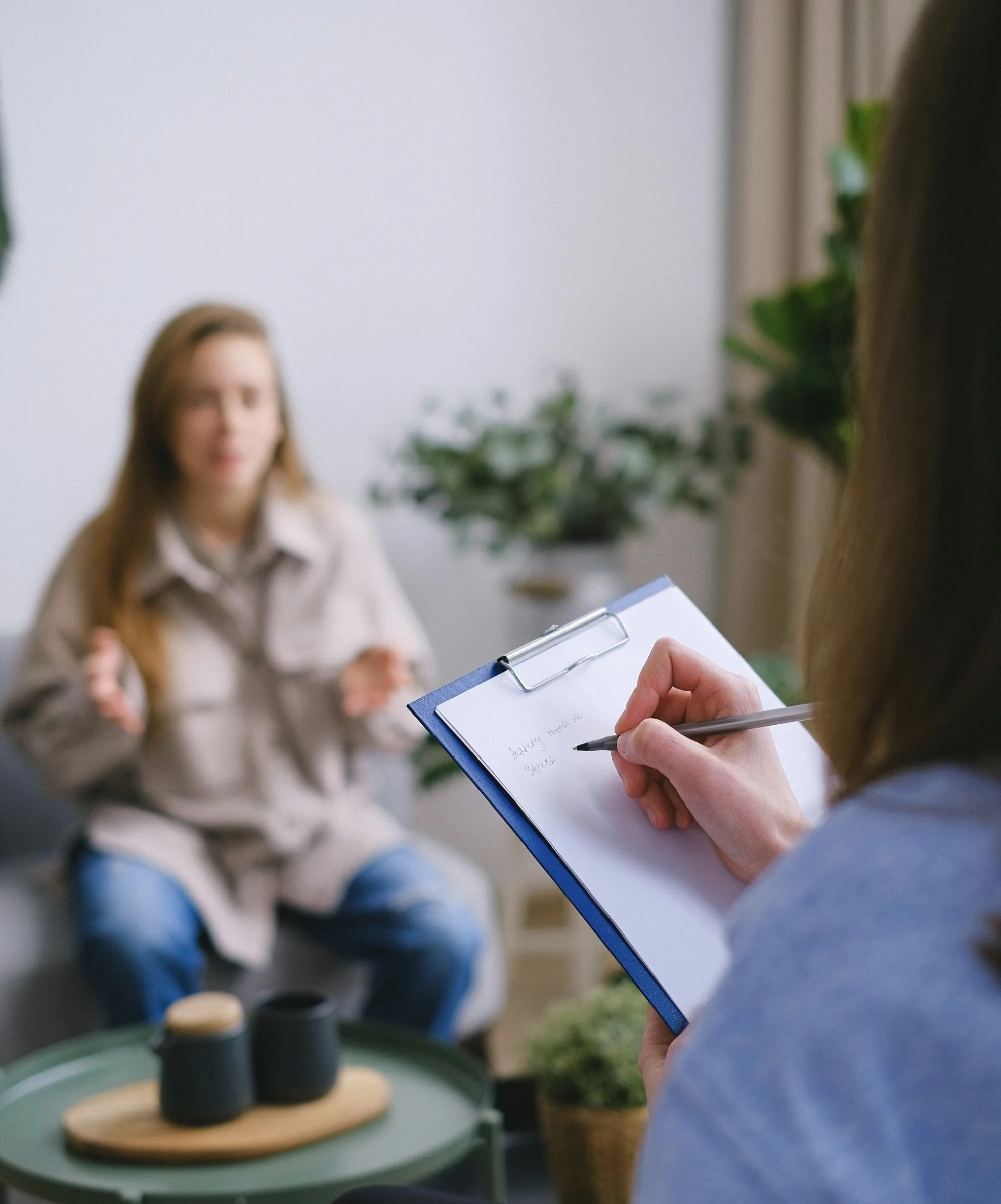 Therapist taking notes during a therapy session with a woman sitting on a couch in a cozy room with plants.
