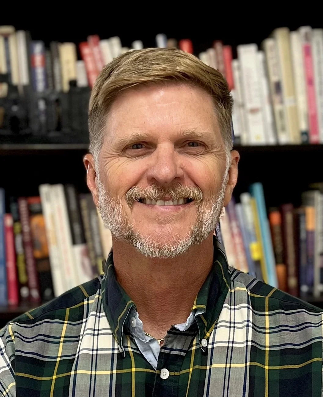 A middle-aged man with a beard and mustache smiling, wearing a plaid shirt, standing in front of a bookshelf filled with books.