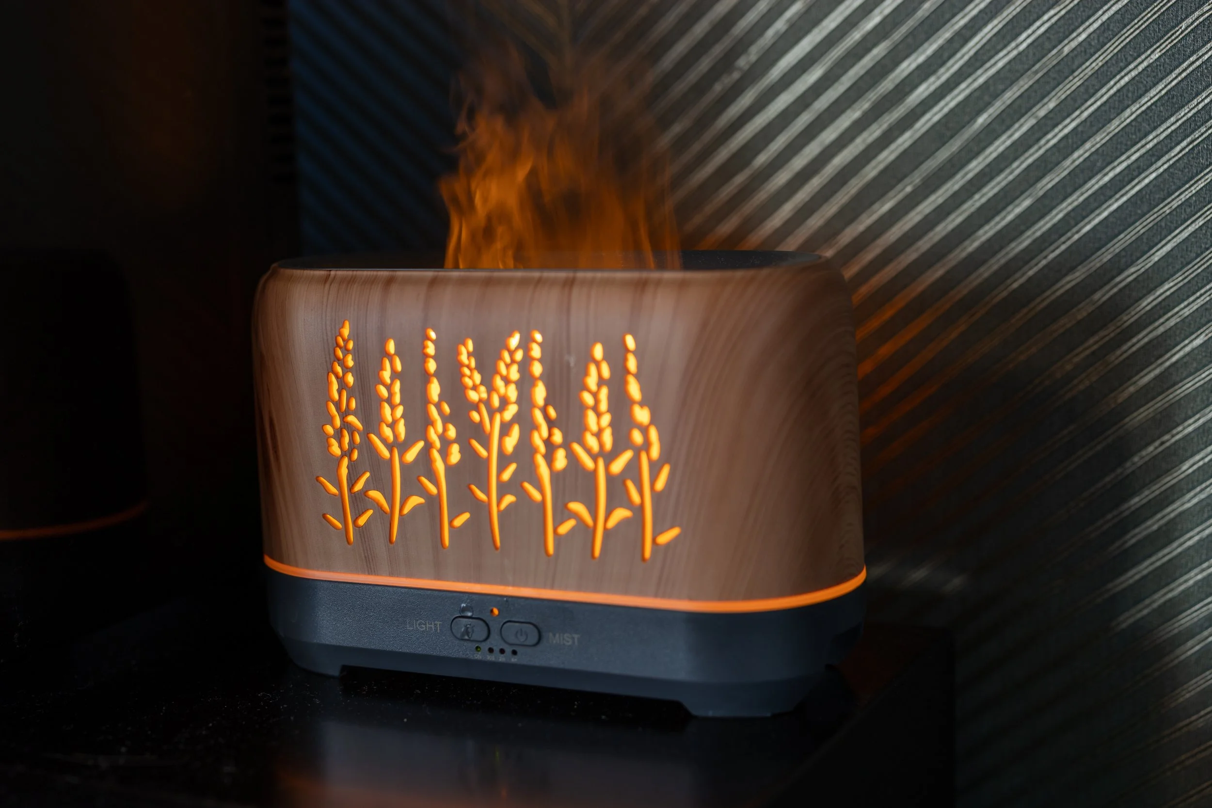 A wooden-patterned diffuser with leaf and stem cutouts emitting orange light, with mist and visible flames inside.