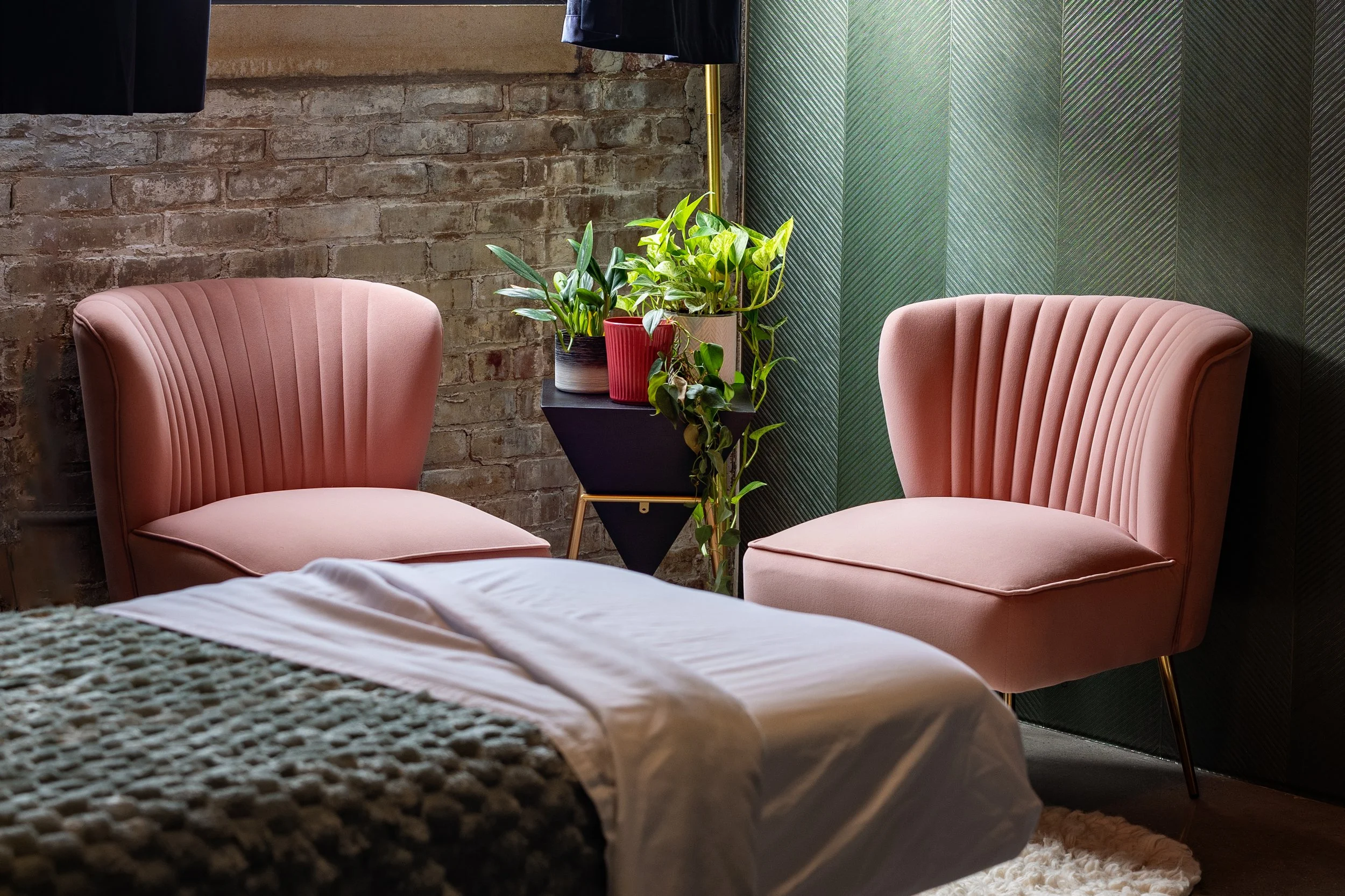 Indoor seating area with two pink velvet armchairs, a bed with white sheets, a gray textured blanket, a small side table with potted plants, and a brick wall with dark curtains.