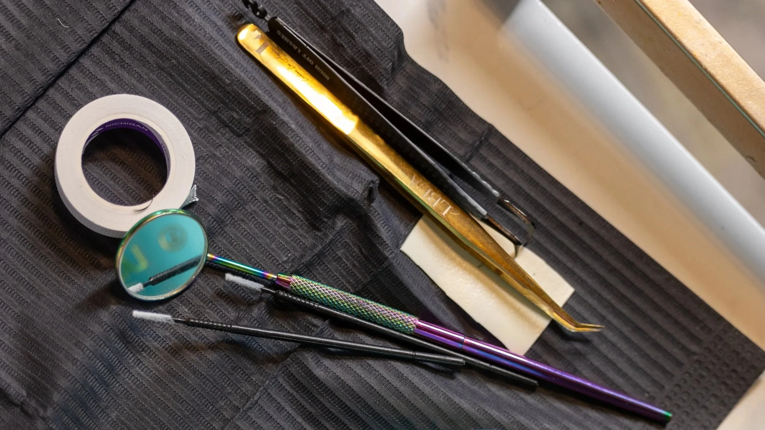 Close-up of a black textured work surface with a roll of white tape, a pair of black tweezers, a colorful tattoo needle with purple and green metallic parts, and a metallic gold tool lying on top.